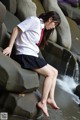 A woman in a school uniform sitting on a rock by the water.